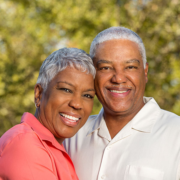 A smiling middle-aged couple posing outdoors, showcasing their dental office-perfected smiles.