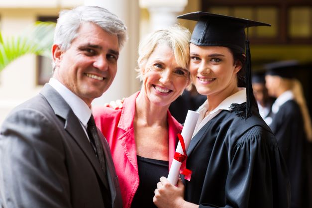 A proud graduate in cap and gown smiling with two older adults, possibly her parents, at a graduation ceremony outside a dental office.
