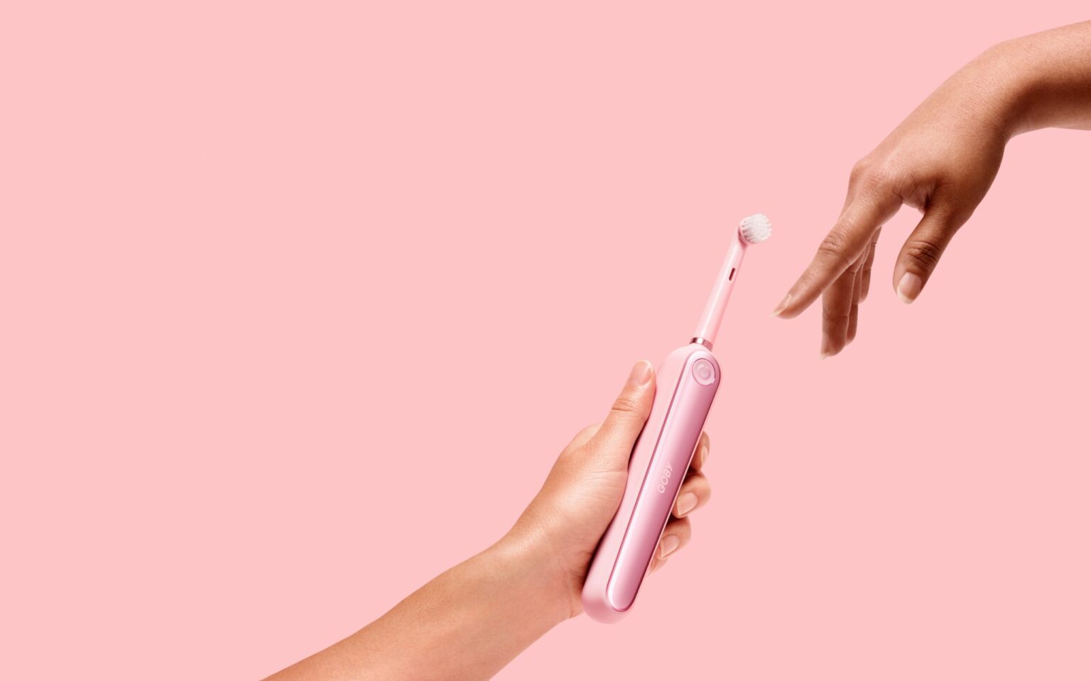 Hand holding an electric toothbrush with another hand applying toothpaste against a pink background, highlighting affordable Dallas dentistry.