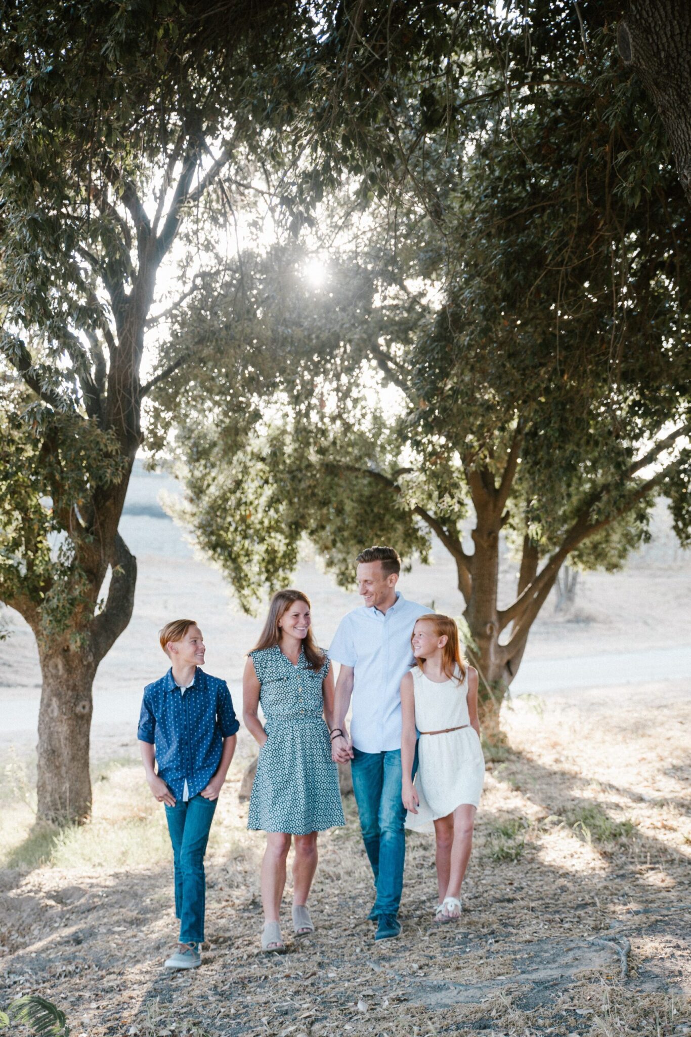A family of four walks hand in hand under the trees, with sunlight filtering through the leaves on their way to an affordable Dallas dentist.