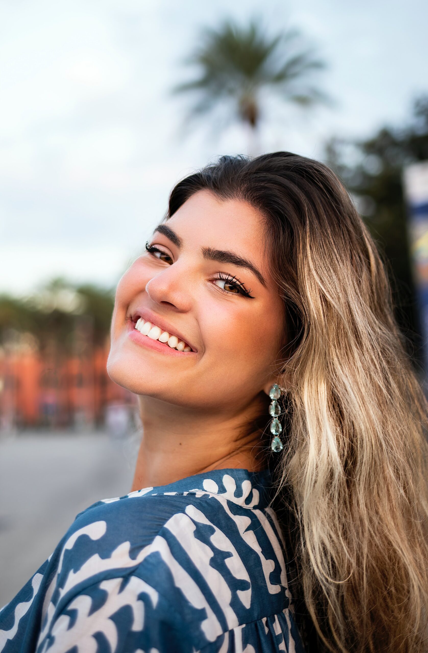 A smiling woman with long blonde hair and earrings, wearing a blue patterned garment, poses in front of a palm tree, capturing the essence of leisure outside a prominent Dallas dentist's office.