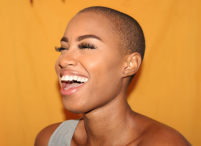 Woman with a shaved head laughing in a dental clinic against a yellow background.