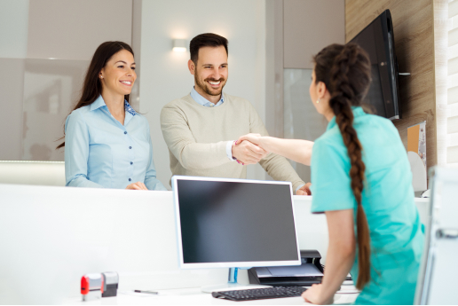 Two professionals shaking hands over a desk in a dental office environment, with another individual looking on.