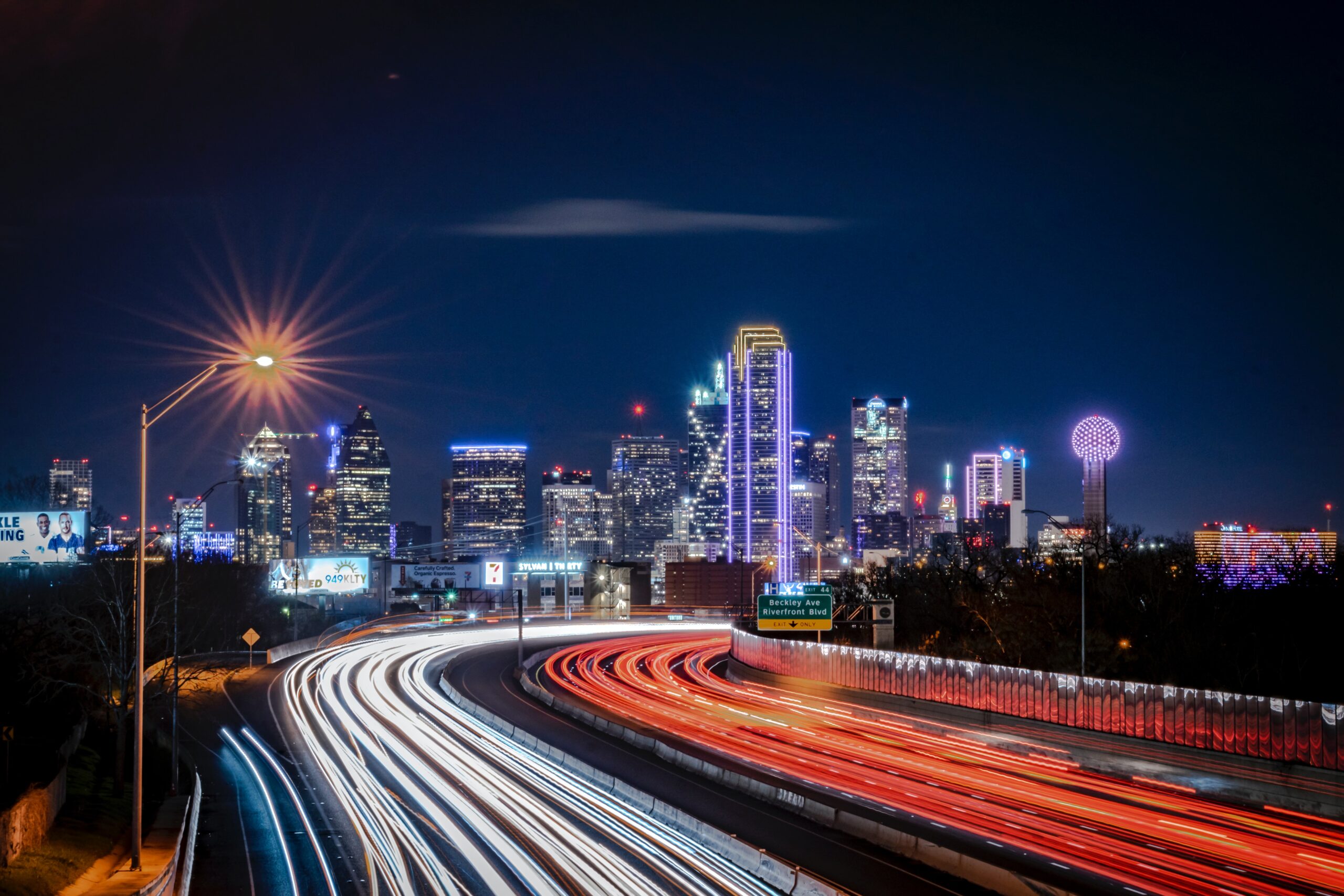 Nighttime view of a city skyline, possibly Dallas, with light trails from vehicular traffic on a highway.