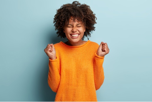 Woman in orange sweater with eyes closed and fists clenched in a celebratory gesture after her visit to an affordable Dallas dentist.
