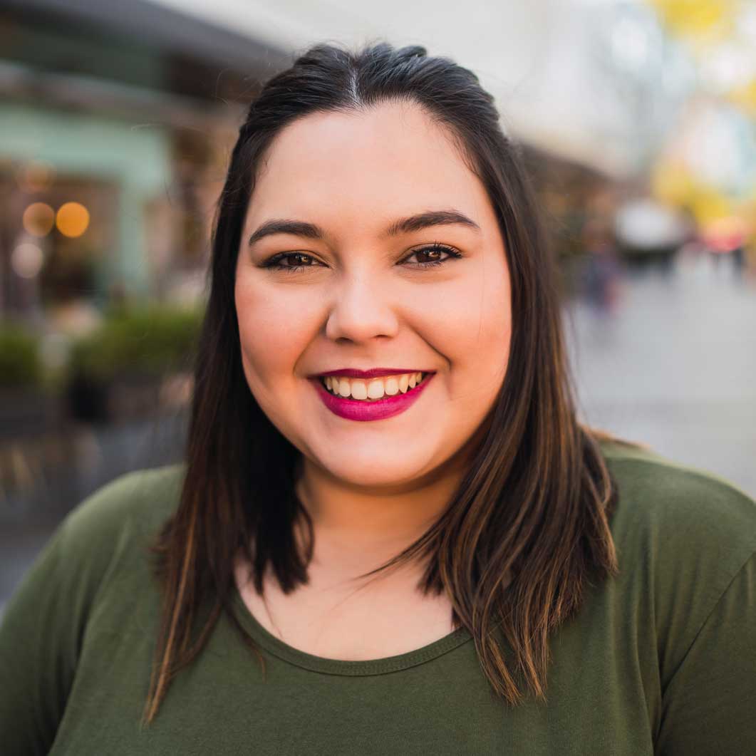 A smiling woman with red lipstick and shoulder-length hair, wearing a green top, stands confidently outside a Dallas dental clinic, with an out-of-focus street scene in the background.