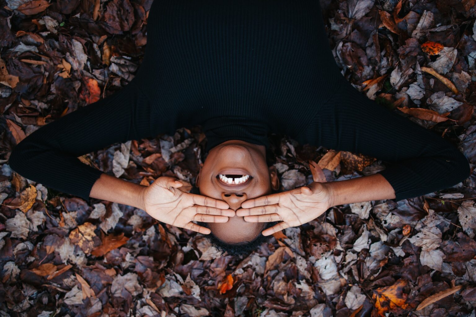 A person lying on a bed of autumn leaves, covering their eyes with their hands and smiling, reminiscent of the carefree joy one feels after an affordable Dallas dentist visit.