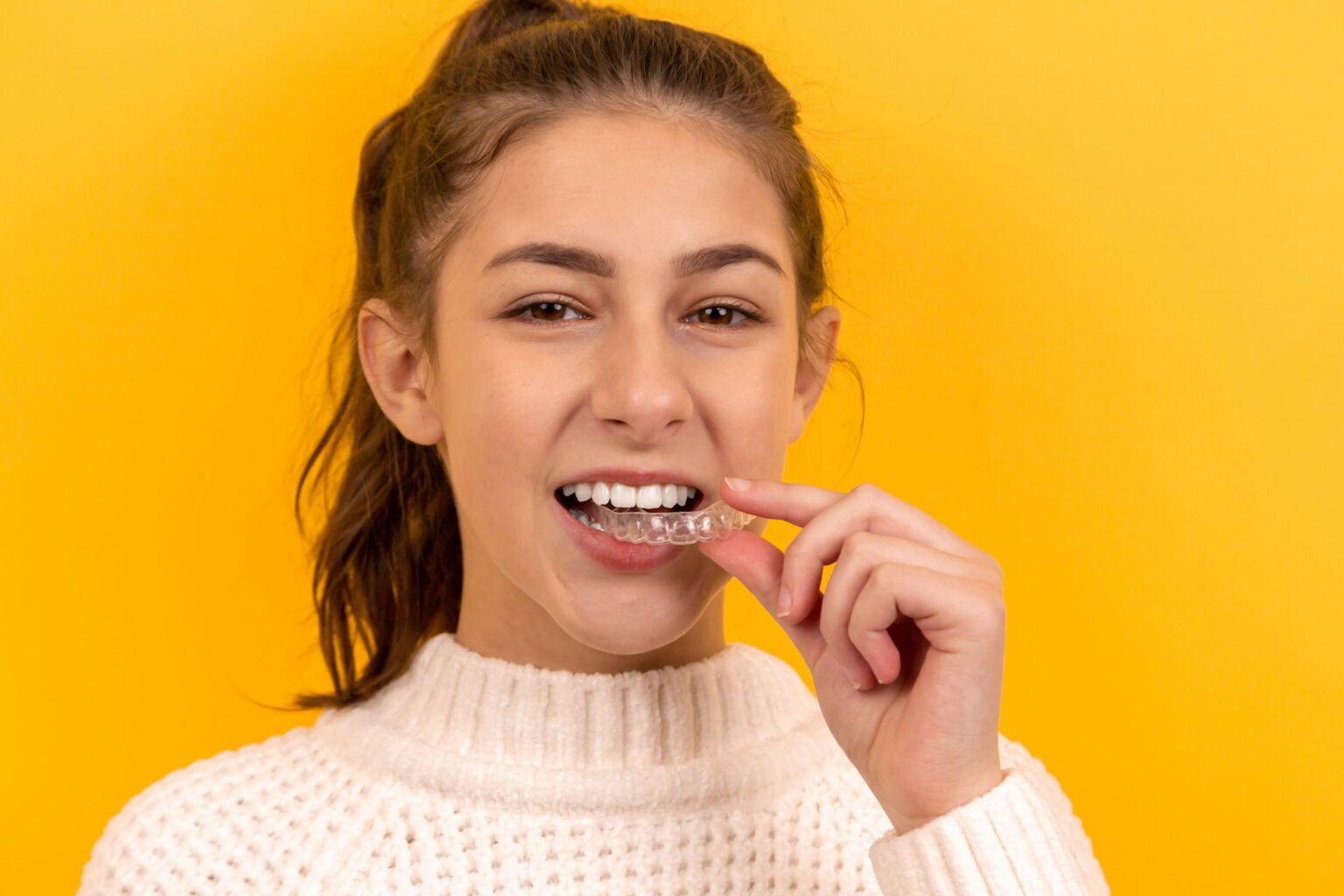 A woman in a white sweater against a yellow background, holding an aligner near her mouth and smiling, showcasing the quality work of an affordable Dallas dentist.