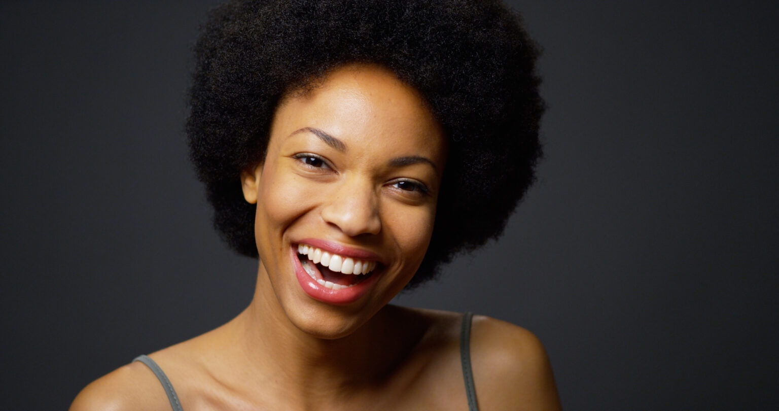 A smiling woman with an afro against a grey background in a dental clinic.