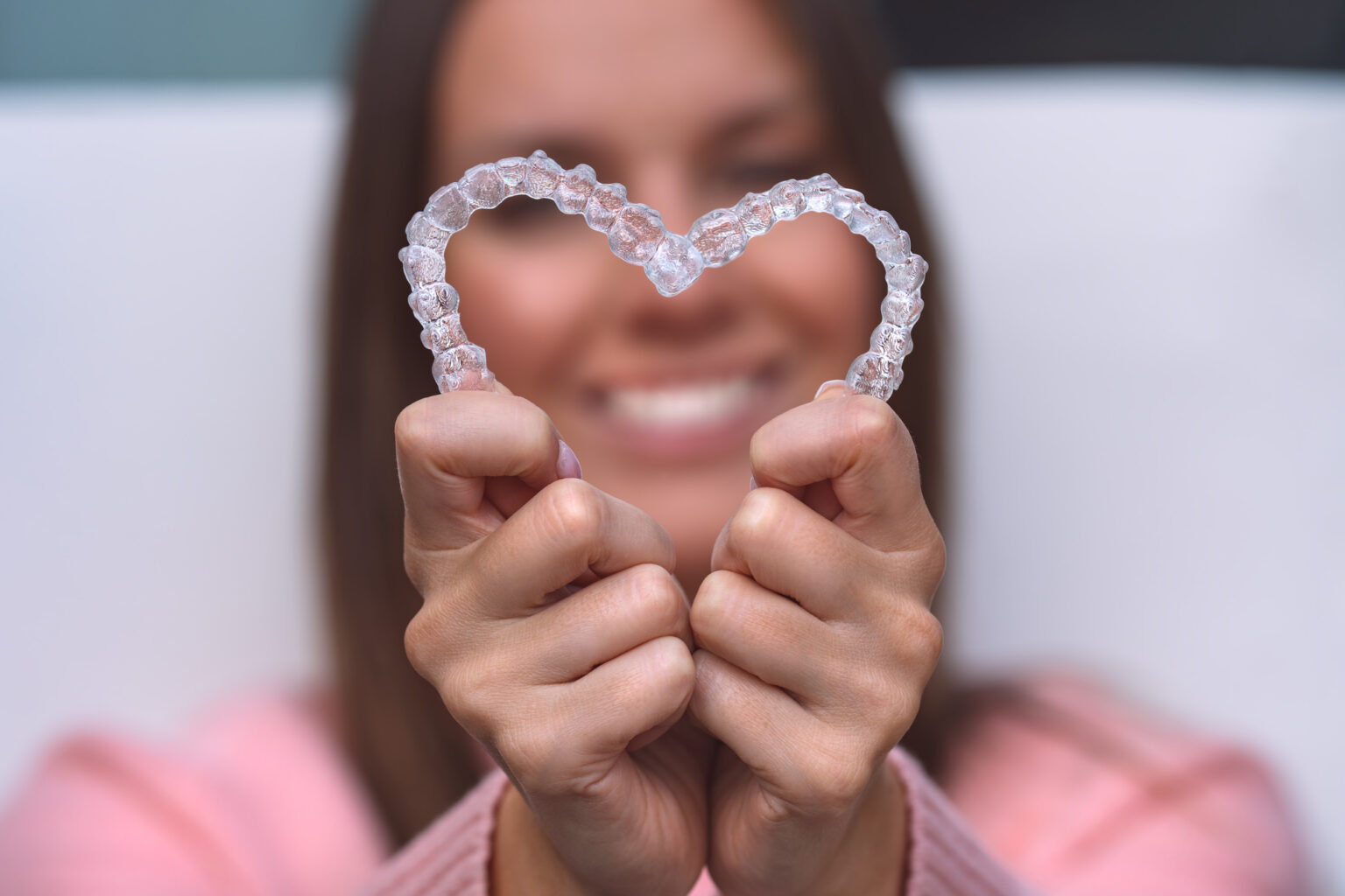 A smiling woman holding up a pair of clear dental aligners in the shape of a heart in a dental clinic.