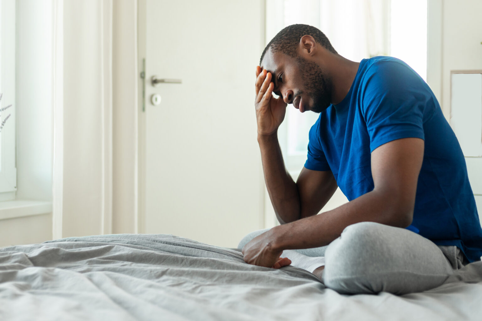 Man sitting on a bed with his hand on his forehead, appearing stressed or upset.