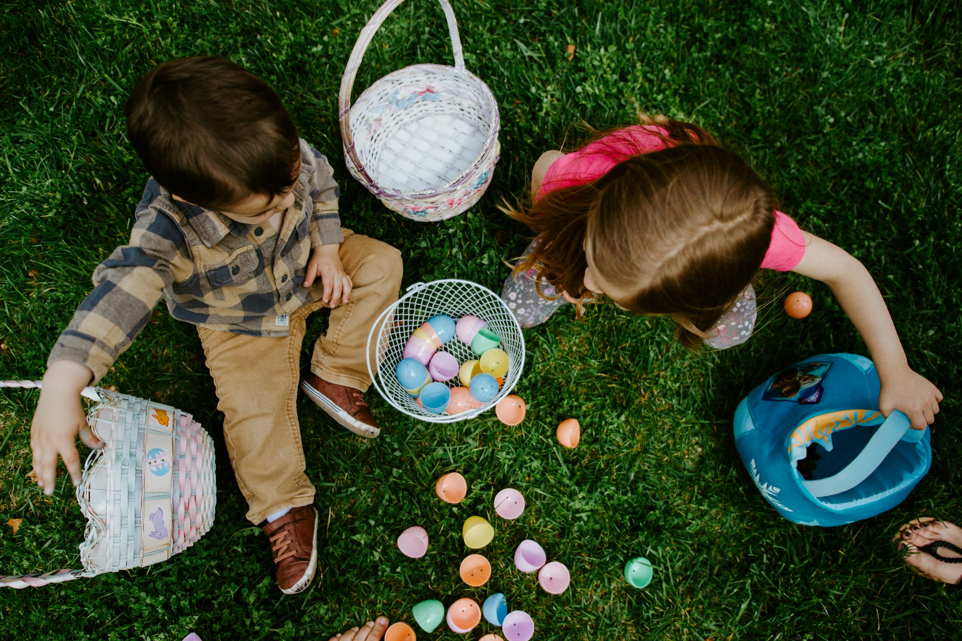 The aftermath of Easter. Two children collecting colorful easter eggs in baskets on a grassy field, viewed from above.