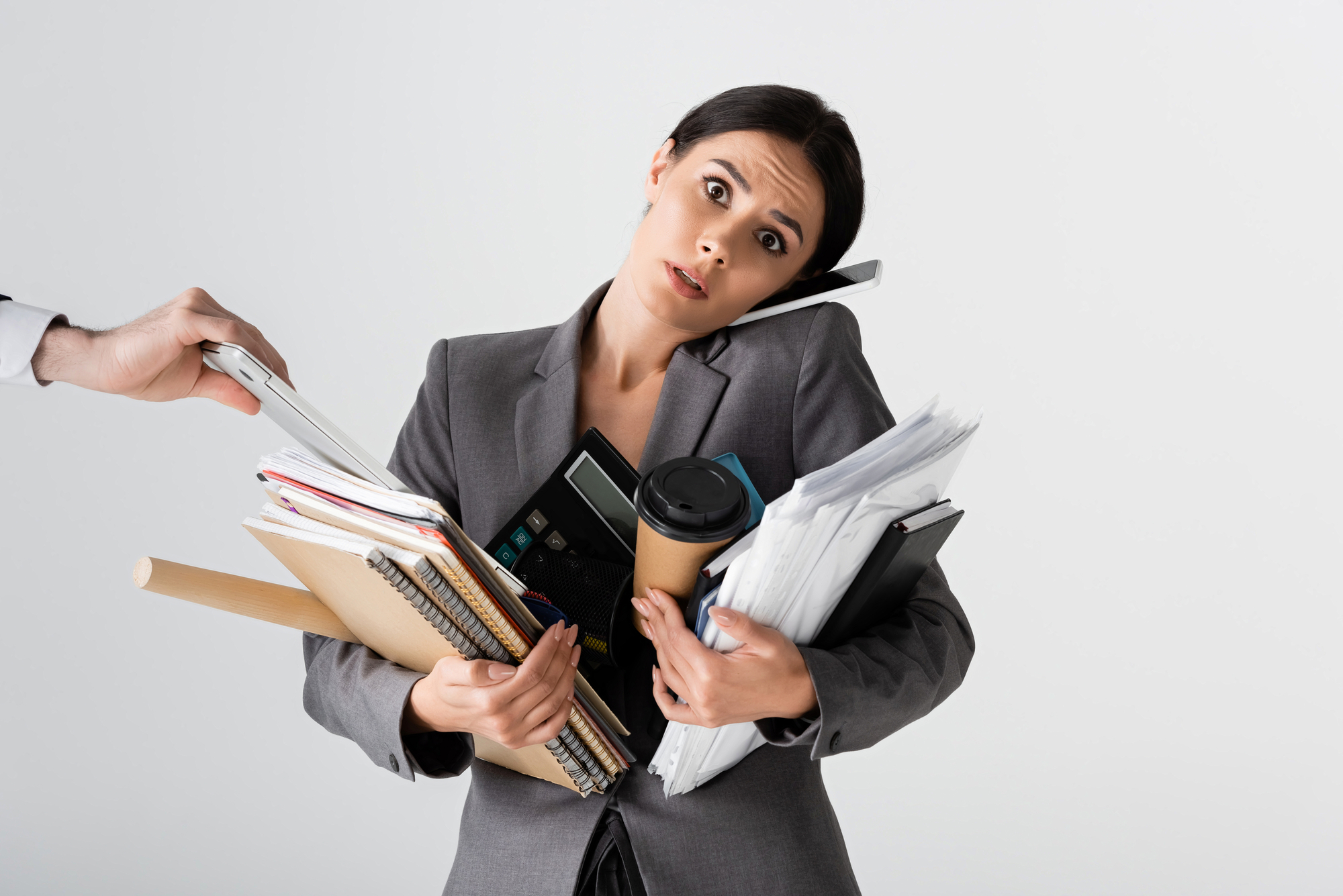 A stressed businesswoman holding documents, coffee, and office supplies as a hand adds more paperwork to her load.