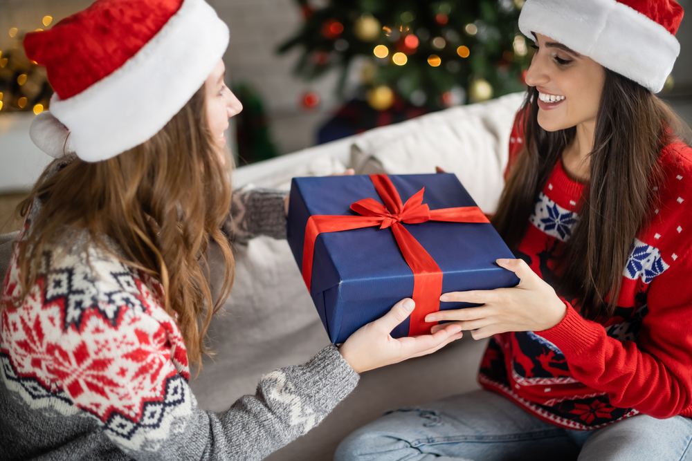Two people wearing Santa hats exchange a gift wrapped in blue paper with a red ribbon. They are seated on a cozy couch, smiling, in a festive room with a decorated Christmas tree in the background.