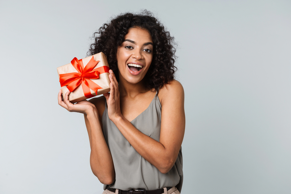A woman with curly hair, wearing a sleeveless top, smiles and holds a gift wrapped in brown paper with a red ribbon. The background is plain light gray.