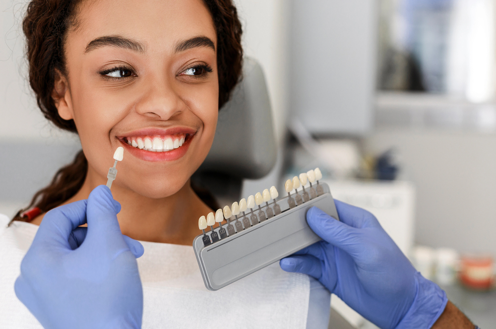 A woman smiles in a dental office as a dentist wearing blue gloves holds a tooth shade guide next to her mouth, matching her teeth color for a possible procedure. She wears a dental bib and appears content.