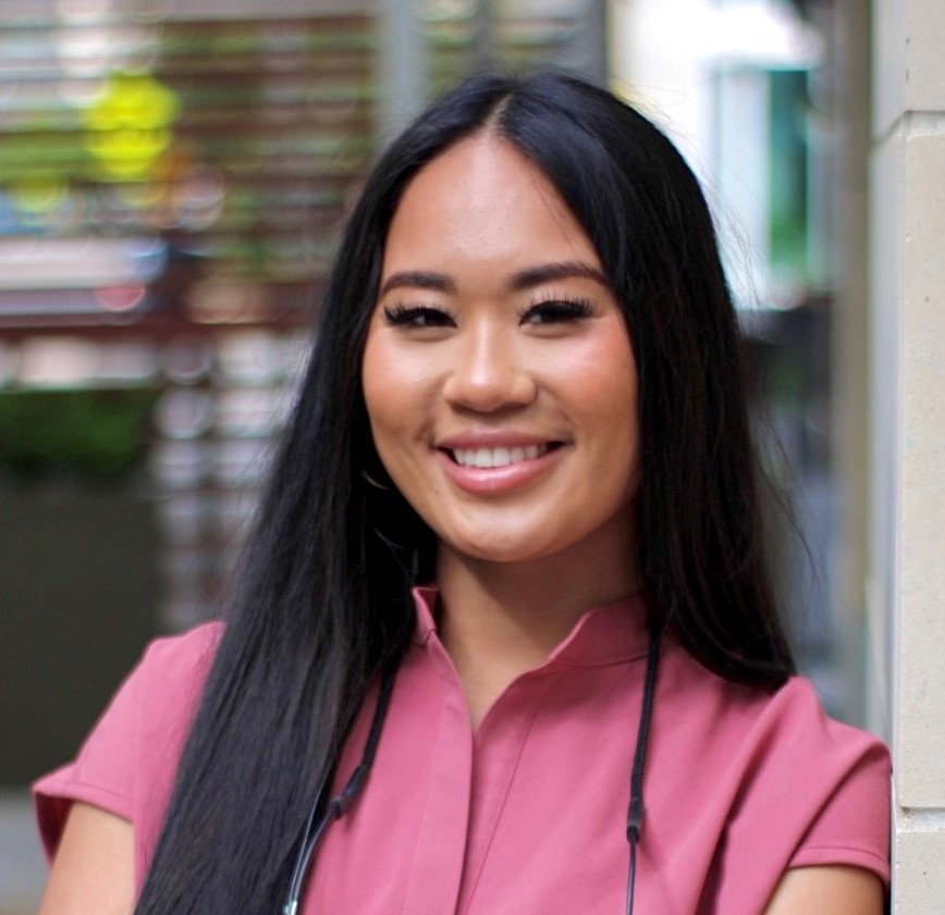 A person with long black hair, wearing a pink top, smiles while standing outdoors. The background is blurred, showing some greenery and buildings. A person with long black hair, wearing a pink top, smiles while standing outdoors. The background is blurred, showing some greenery and buildings.