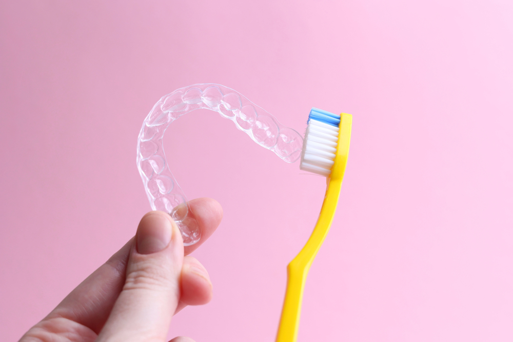 A person holds a clear Invisalign aligner in one hand and a yellow toothbrush in the other, against a pink background.