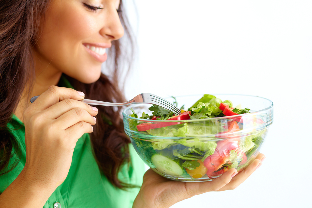 A woman in a green shirt smiles while holding a fork and a bowl of salad containing lettuce, cucumbers, tomatoes, and other vegetables. The background is a plain white, emphasizing the fresh ingredients.