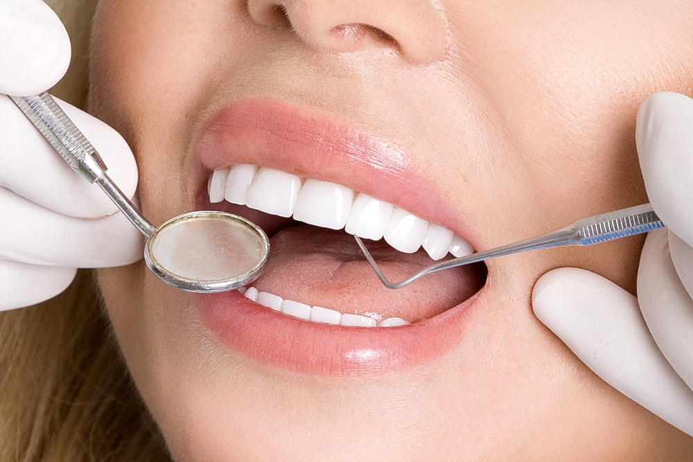 A close-up of a person at a dental check-up, with a dentist's gloved hands holding dental tools. The person's mouth is open, showing clean, white teeth, as the dentist examines them with a mirror and probe.