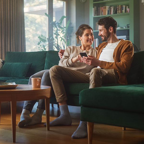A couple sits closely together on a green sofa in a cozy living room, smiling as they look at a smartphone—possibly booking a dentist appointment. Sunlight streams through large windows, and a mug rests on the wooden coffee table in front of them. A couple sits closely together on a green sofa in a cozy living room, smiling as they look at a smartphone—possibly booking a dentist appointment. Sunlight streams through large windows, and a mug rests on the wooden coffee table in front of them.