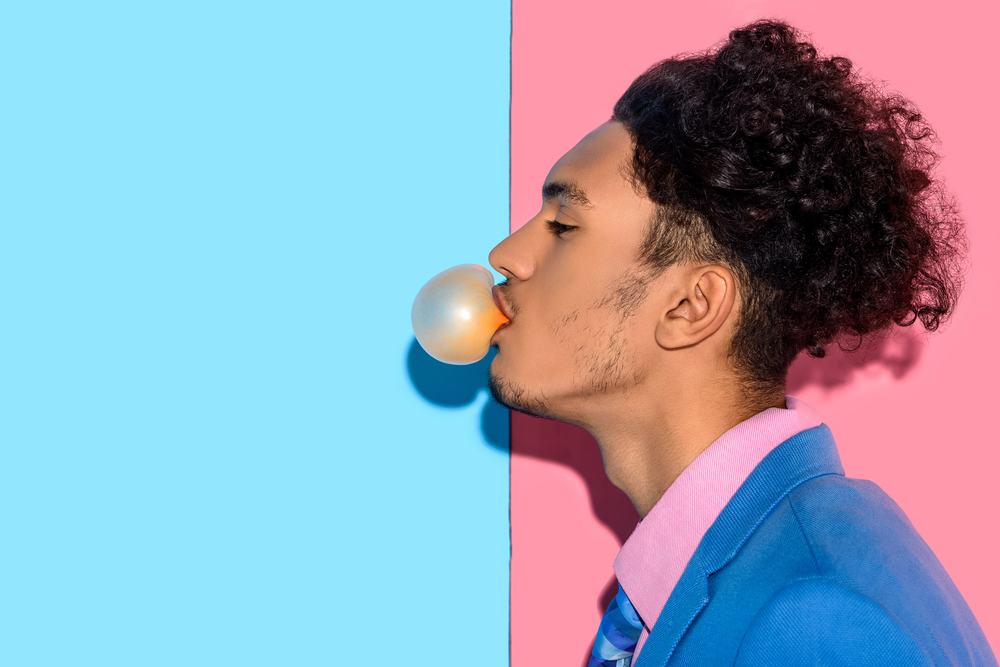 A young man in a blue suit and pink shirt blows a bubble with gum. He is in profile against a split background, with one half blue and the other half pink.