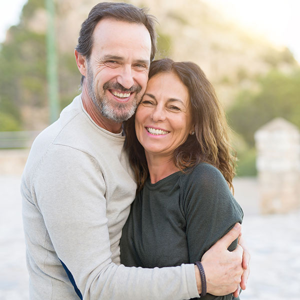 A smiling middle-aged couple stands outdoors, embracing each other warmly. The background is softly blurred, showing sunlight and greenery.
