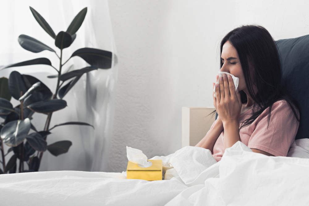 A woman sits up in bed, holding a tissue to her nose as if sneezing or blowing her nose. A yellow tissue box sits nearby on the bed. There is a large plant in the background.