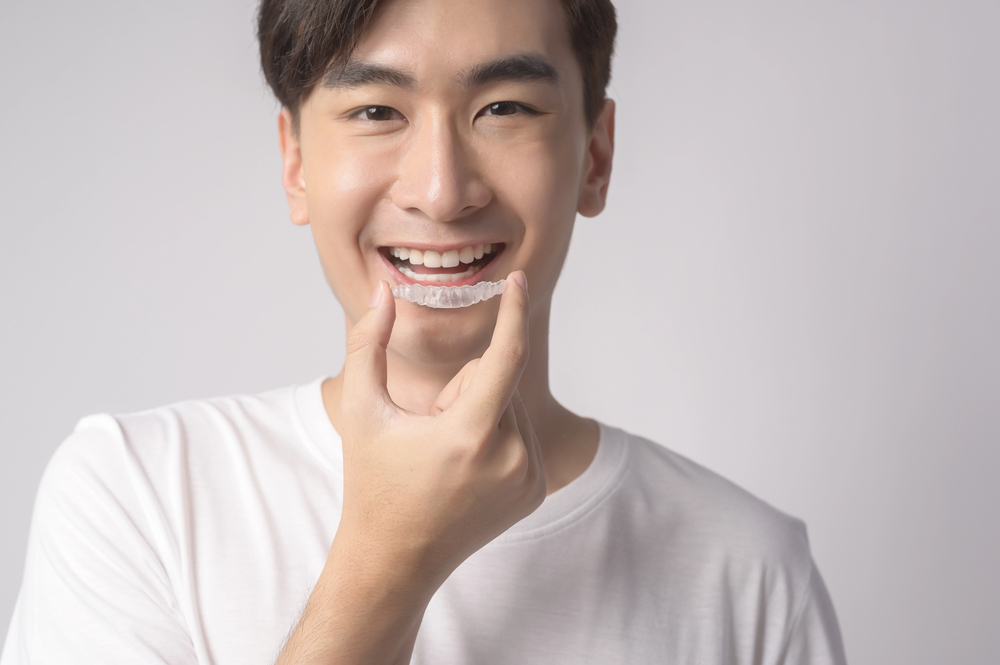 A young man in a white shirt smiles while holding a clear dental aligner in front of his mouth against a plain light background.
