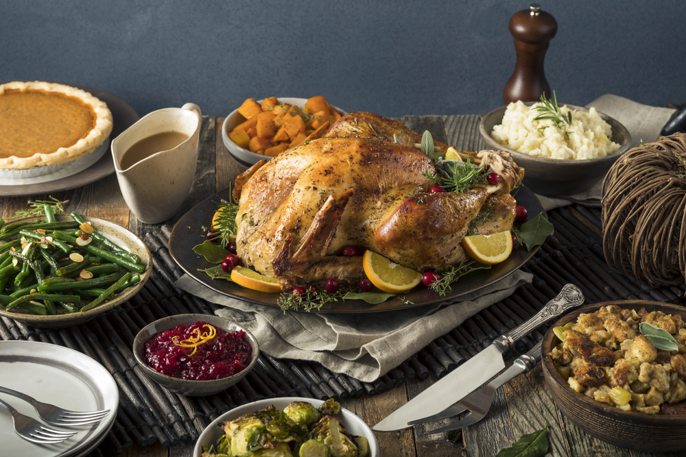 A roasted turkey garnished with herbs and lemon slices is surrounded by classic Thanksgiving dishes, including mashed potatoes, stuffing, green beans, cranberry sauce, sweet potatoes, Brussels sprouts, gravy, and pumpkin pie.
