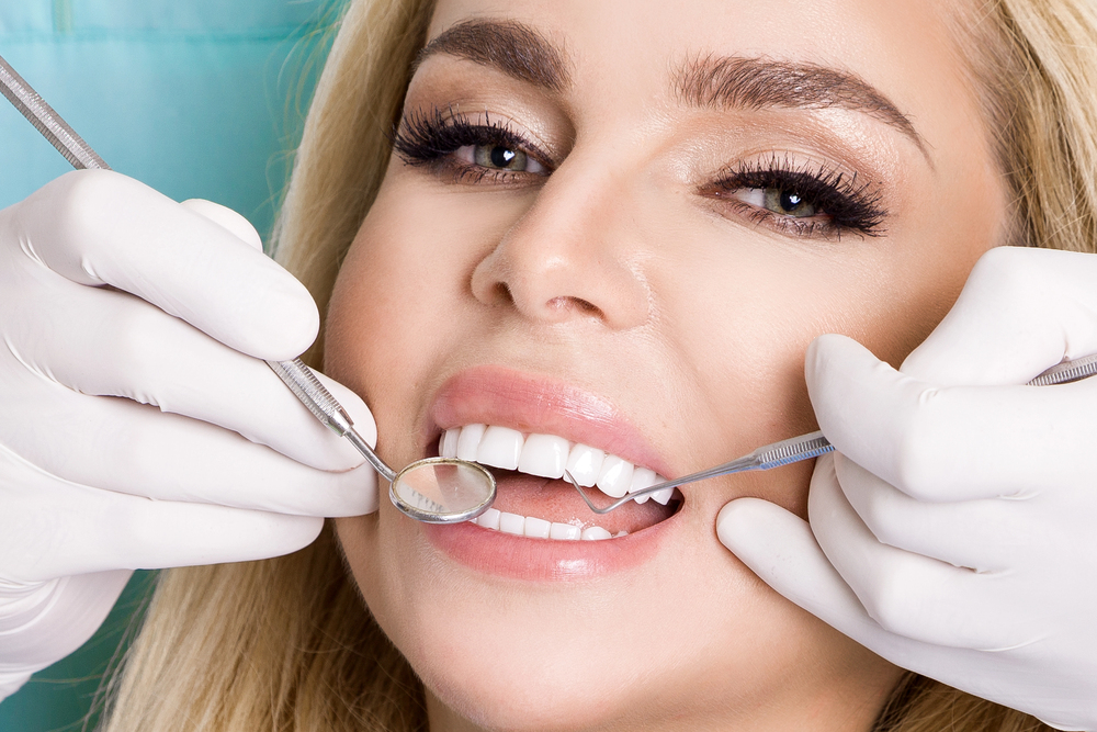 A close-up of a woman smiling during a dental check-up. She has bright white teeth, possibly enhanced by veneers, and a dentist wearing white gloves is holding dental instruments to examine her mouth.