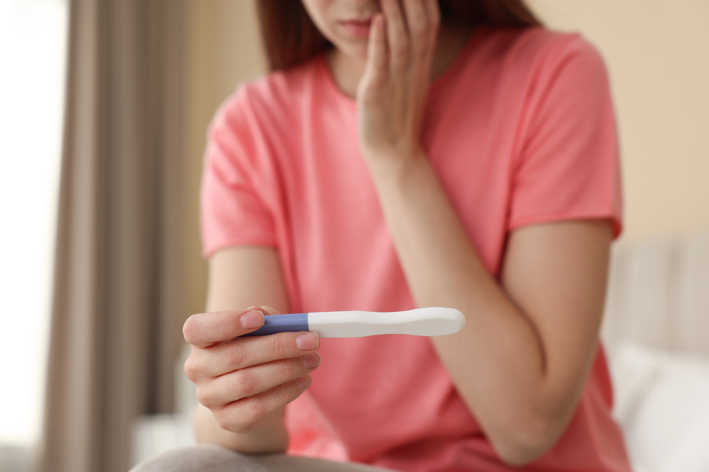 A woman in a pink shirt sits and looks at a pregnancy test, holding her other hand to her face with a concerned expression, similar to waiting anxiously for news from a dentist in Dallas TX. The test result is not visible.