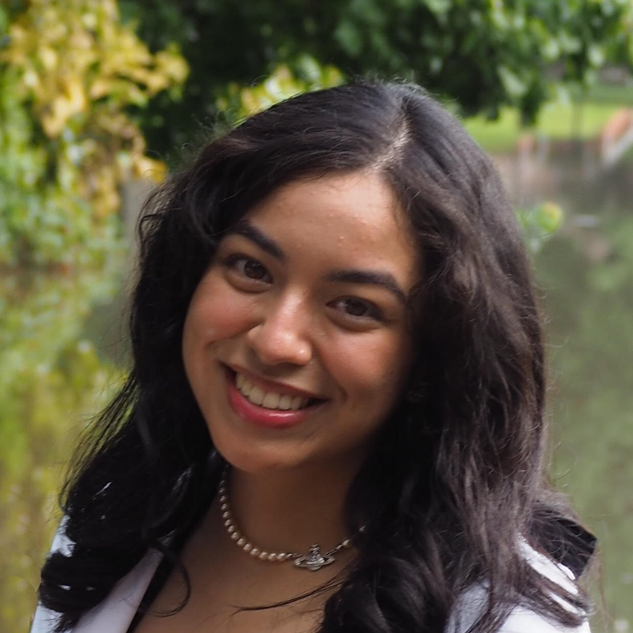 A young woman with long, dark wavy hair smiles at the camera. She is wearing a white top, a pearl necklace, and is outdoors with green trees and a body of water blurred in the background.