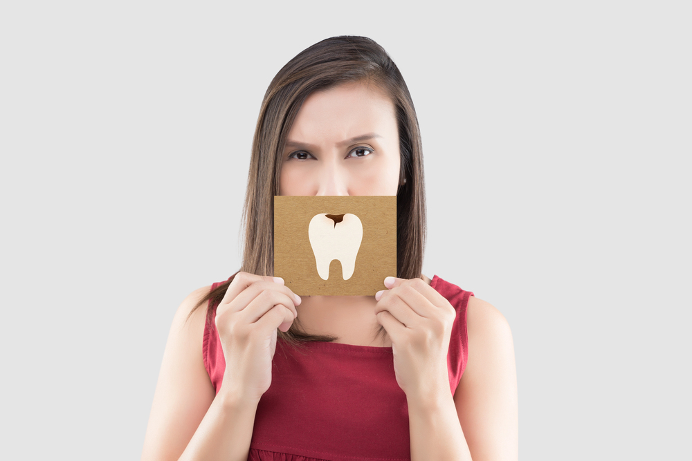 A woman in a red sleeveless top holds a brown card with a drawing of a tooth showing a cavity in front of her mouth, against a plain light gray background.