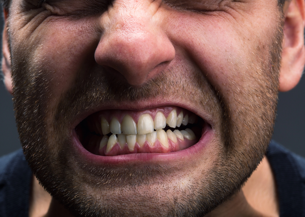 Close-up of a man's face showing him clenching his teeth tightly, eyes shut, and brow furrowed, suggesting tension or stress.