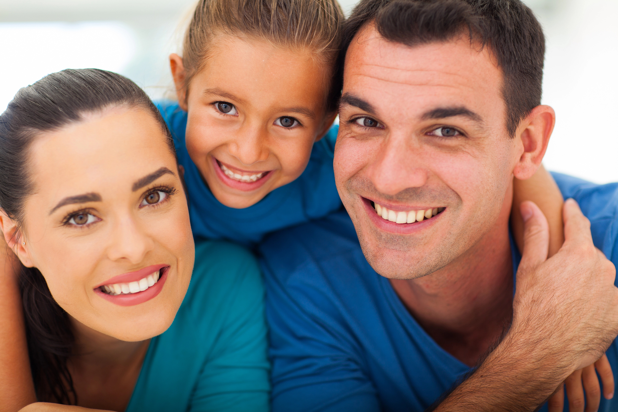 A smiling family of three—a woman, a young girl, and a man—pose closely together in blue tops. The girl is in the middle, hugging both adults, as they all look at the camera, showcasing their happy smiles after visiting a Dentist in Dallas.