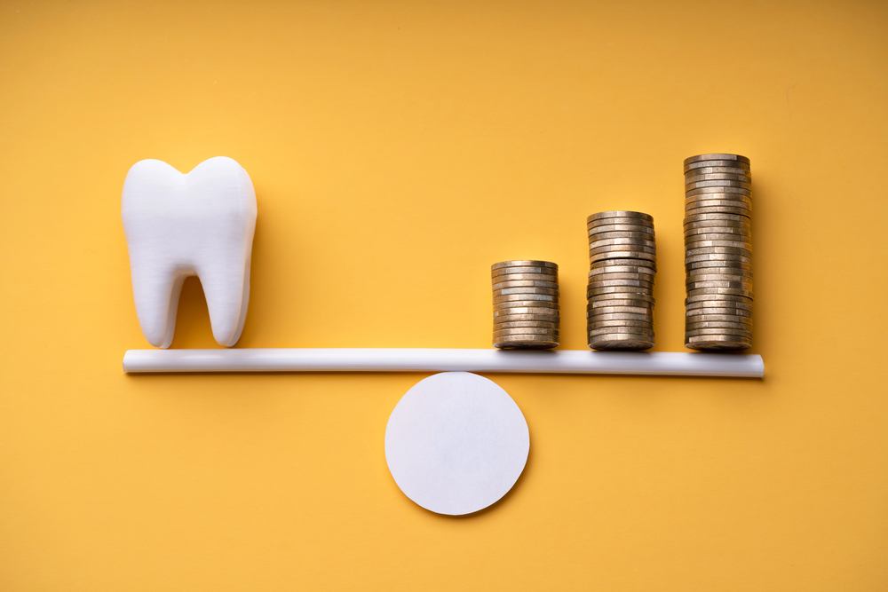 A model tooth balances on one end of a seesaw opposite three stacks of coins, set on a yellow background, illustrating the cost of dental care.