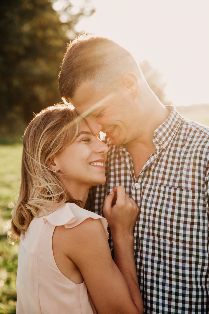 A smiling couple embraces outdoors in warm sunlight. The woman rests her head on the man's chest, both appear happy and relaxed, with trees and grass softly blurred in the background.